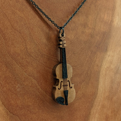 Wooden necklace pendant created in the shape of a miniature violin. Made from hard maple wood and hanging from a black stainless steel chain against a cherry wood background.