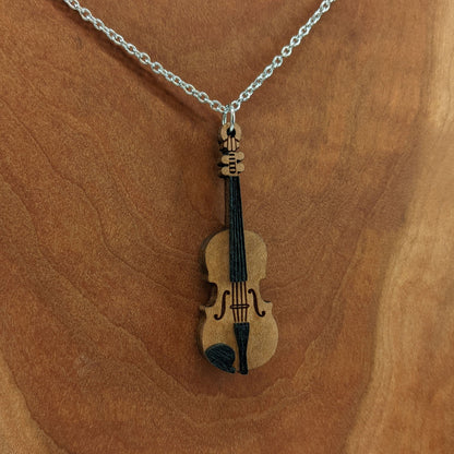 Wooden necklace pendant created in the shape of a miniature violin. Made from hard maple wood and hanging from a silver stainless steel chain against a cherry wood background.