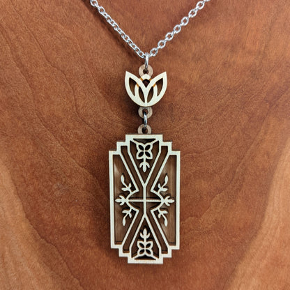 Wooden necklace pendant made from two contrasting layers of birch wood. The top lighter layer is intricately cut with flower like details. The overall shape is rectangular. It hangs from a silver stainless steel chain against a cherry background.