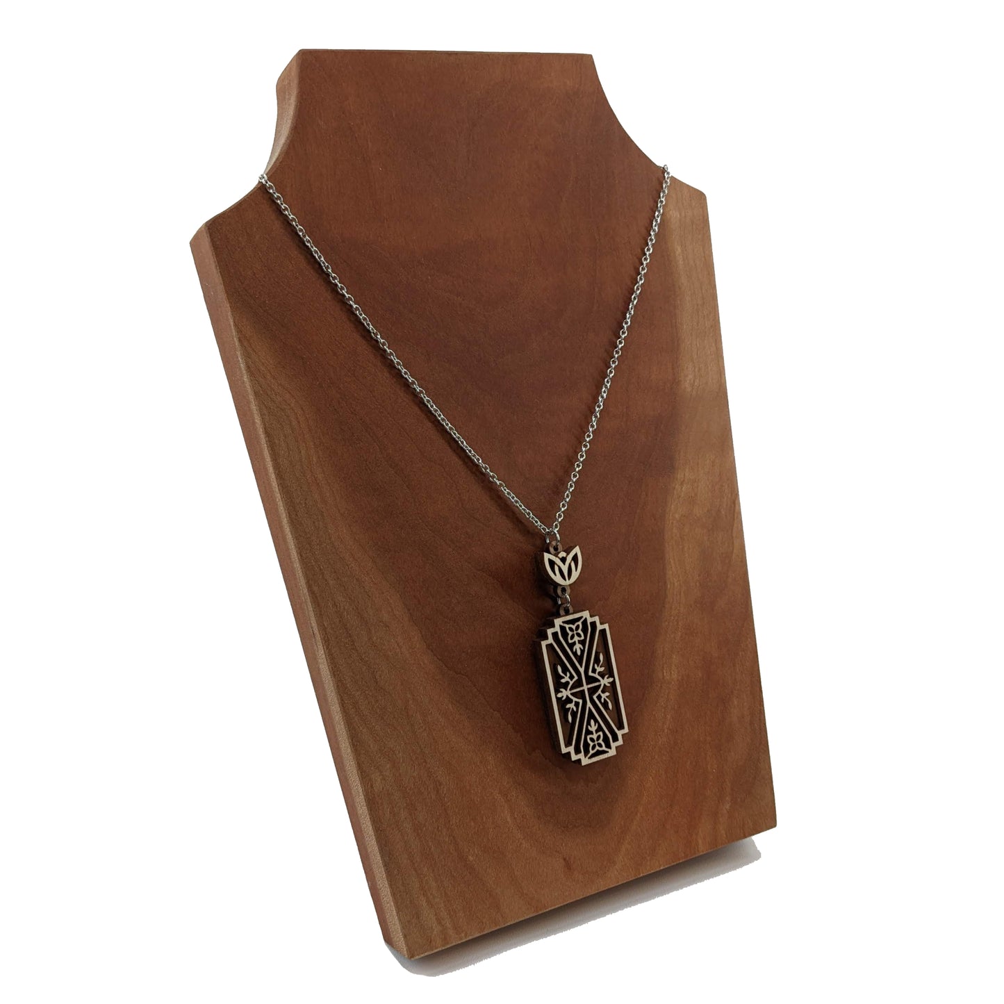 Wooden necklace pendant made from two contrasting layers of birch wood. The top lighter layer is intricately cut with flower like details. The overall shape is rectangular. It hangs from a silver stainless steel chain against a cherry background.