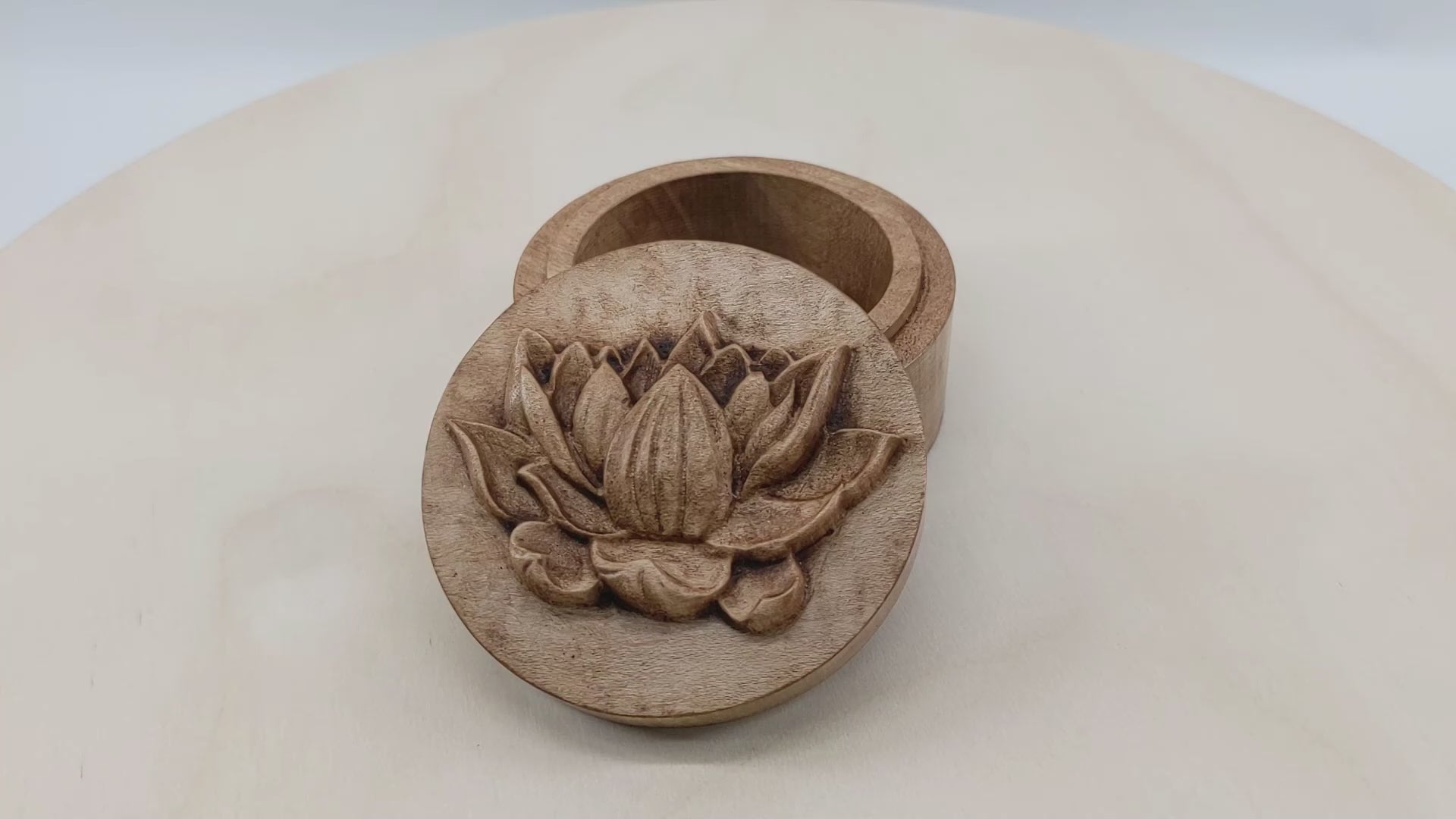 Round wooden box 3D carved with a side profile of a lotus flower. It has several layers of leaves closer to the bottom and layers of petals going up the flower to the pointed center. Made from hard maple wood stained brown against a white background rotating on a table.