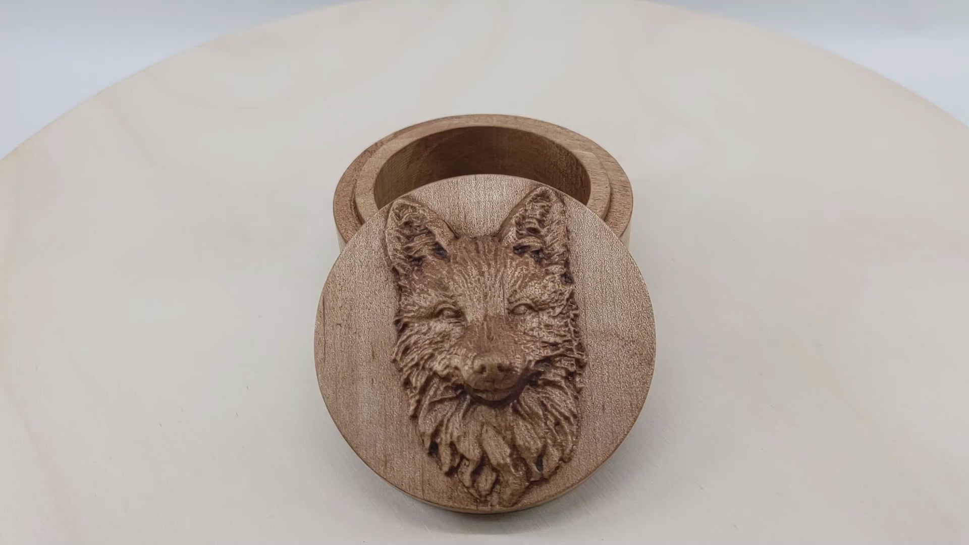 Round wooden box 3D carved with a fox face looking straight ahead. It has tall upright ears and a narrow face. Its fur is long and bushy around its mane and short near its nose. Made from hard maple wood with a brown stain against a white background rotating on a table.