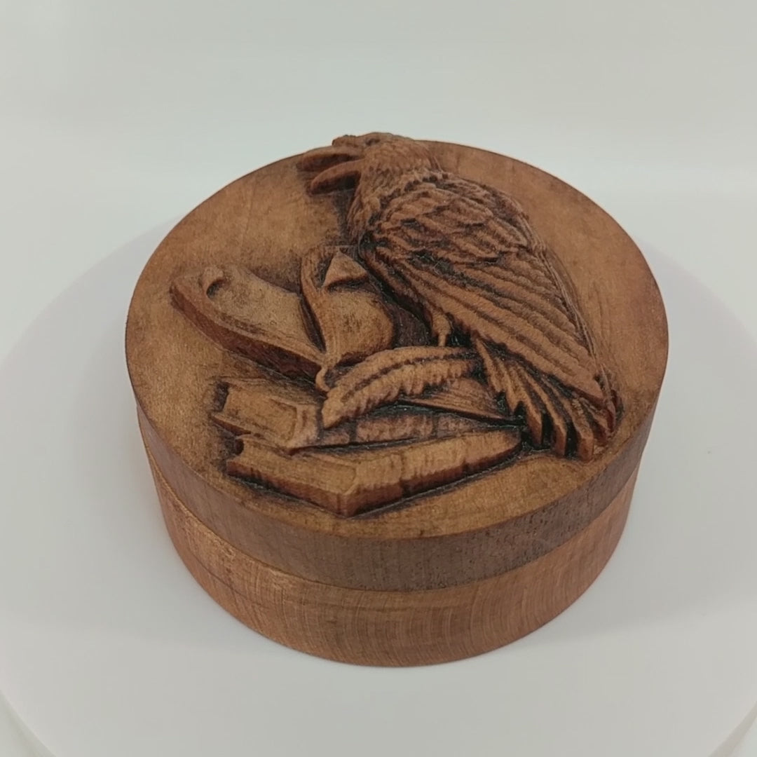 Round wooden box 3D carved with a raven standing three books with a quill at its feet, the top book open and pages turning. The raven has an open beak and intricate layered feathers Made from hard maple wood stained brown against a white background rotating on a table.