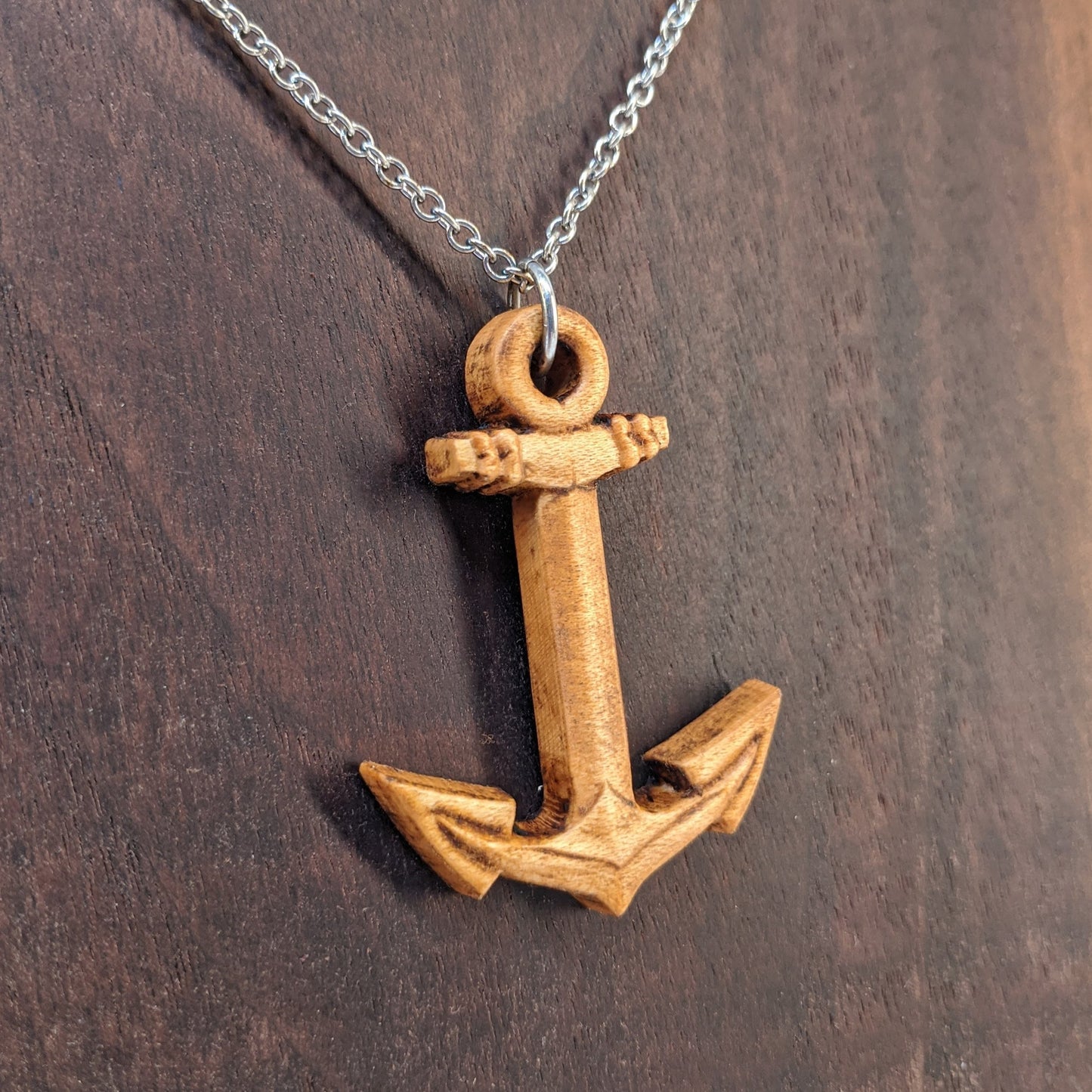 Wooden pendant necklace carved in the shape of a anchor. Made from hard maple wood. It hangs from a silver stainless steel chain against a dark walnut background.