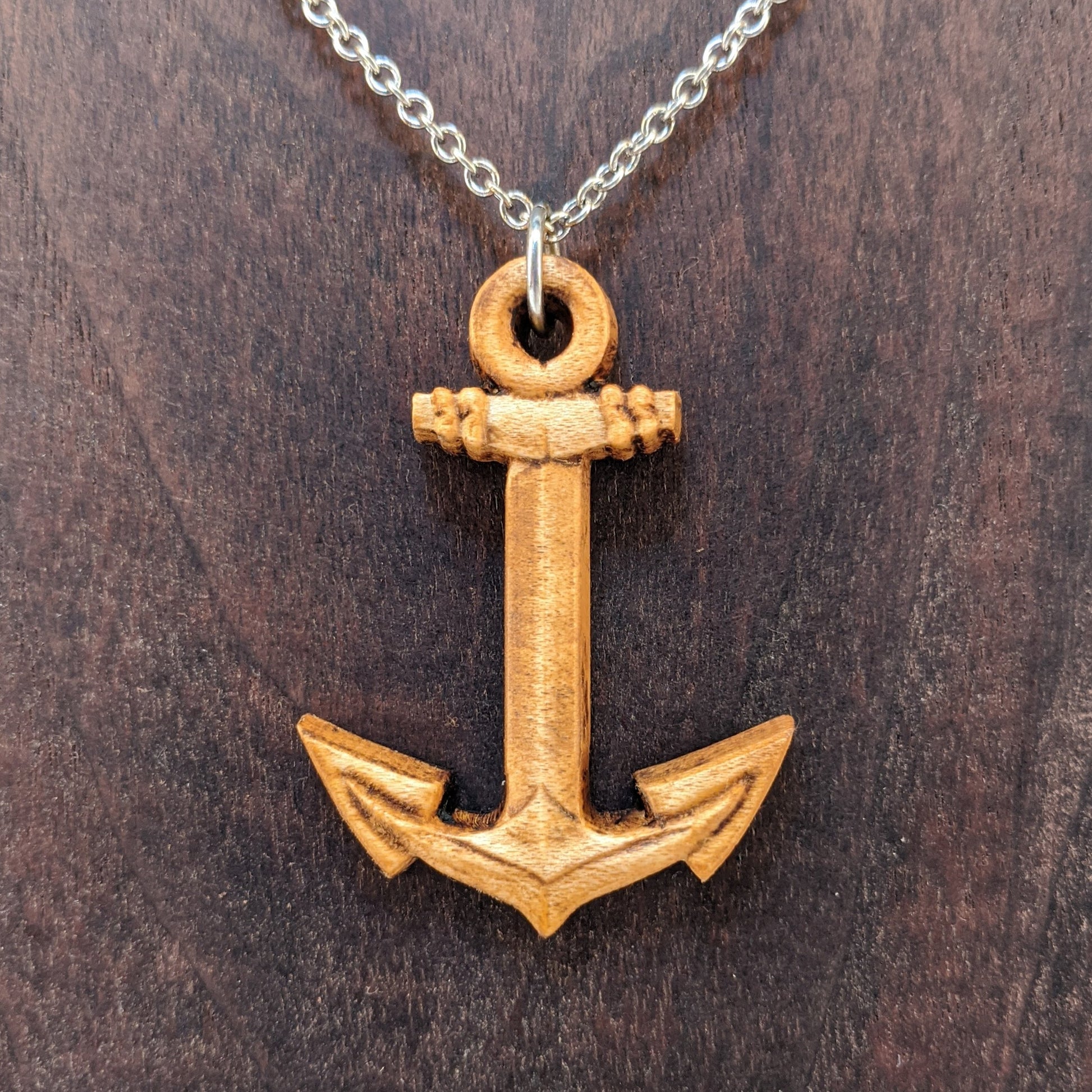 Wooden pendant necklace carved in the shape of a anchor. Made from hard maple wood. It hangs from a silver stainless steel chain against a dark walnut background.