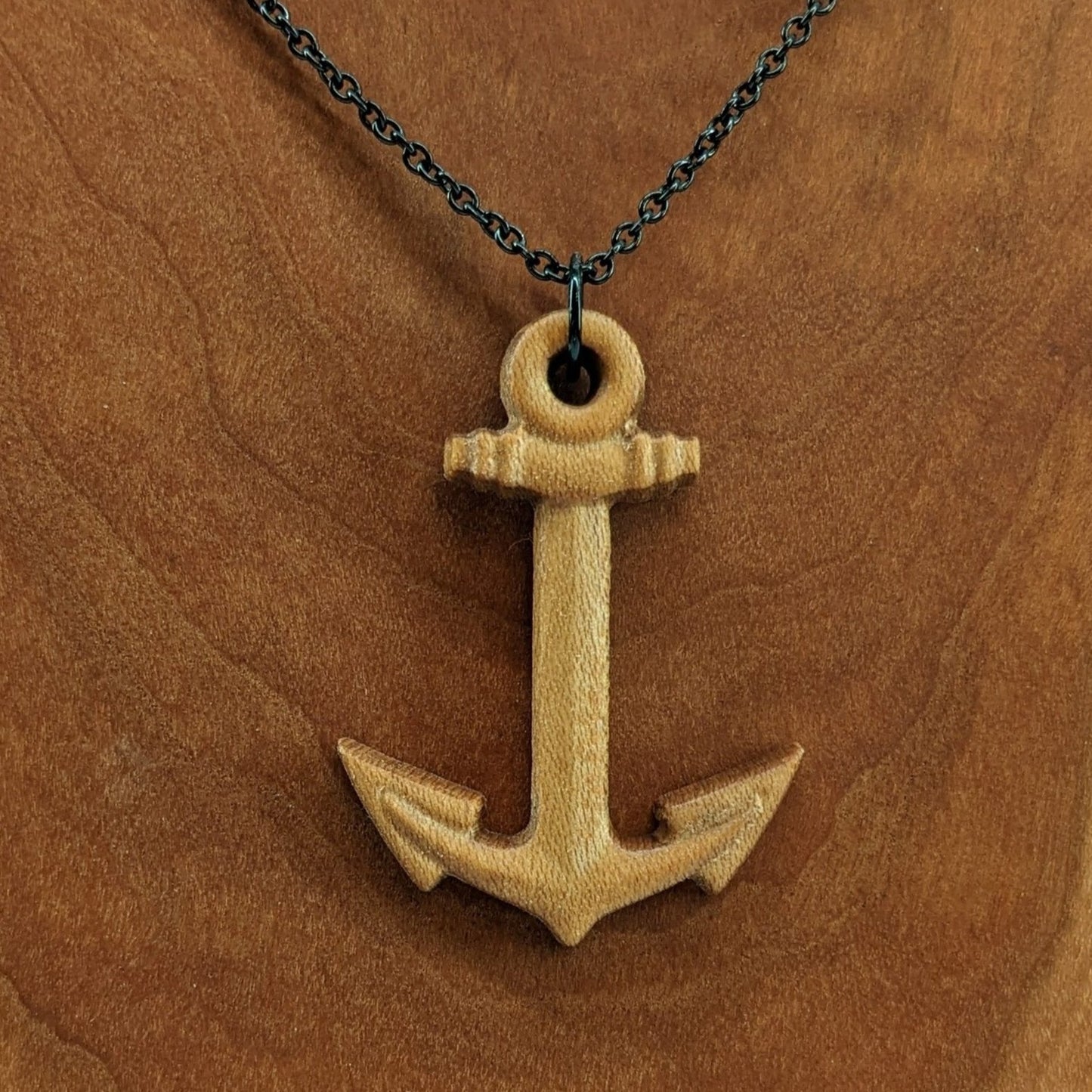 Wooden pendant necklace carved in the shape of a anchor. Made from hard maple wood. It hangs from a black stainless steel chain against a cherry wood background.