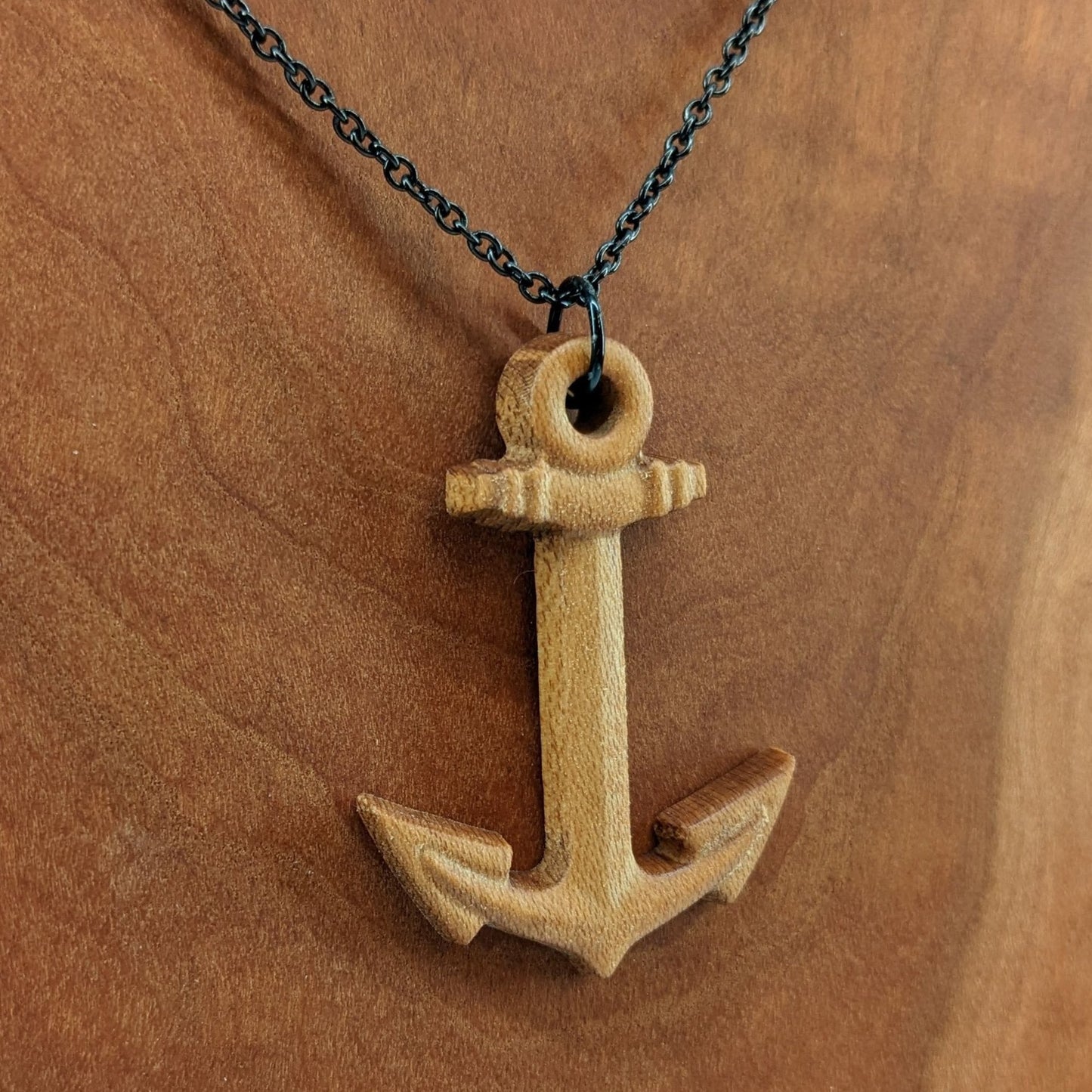 Wooden pendant necklace carved in the shape of a anchor. Made from hard maple wood. It hangs from a black stainless steel chain against a cherry wood background.