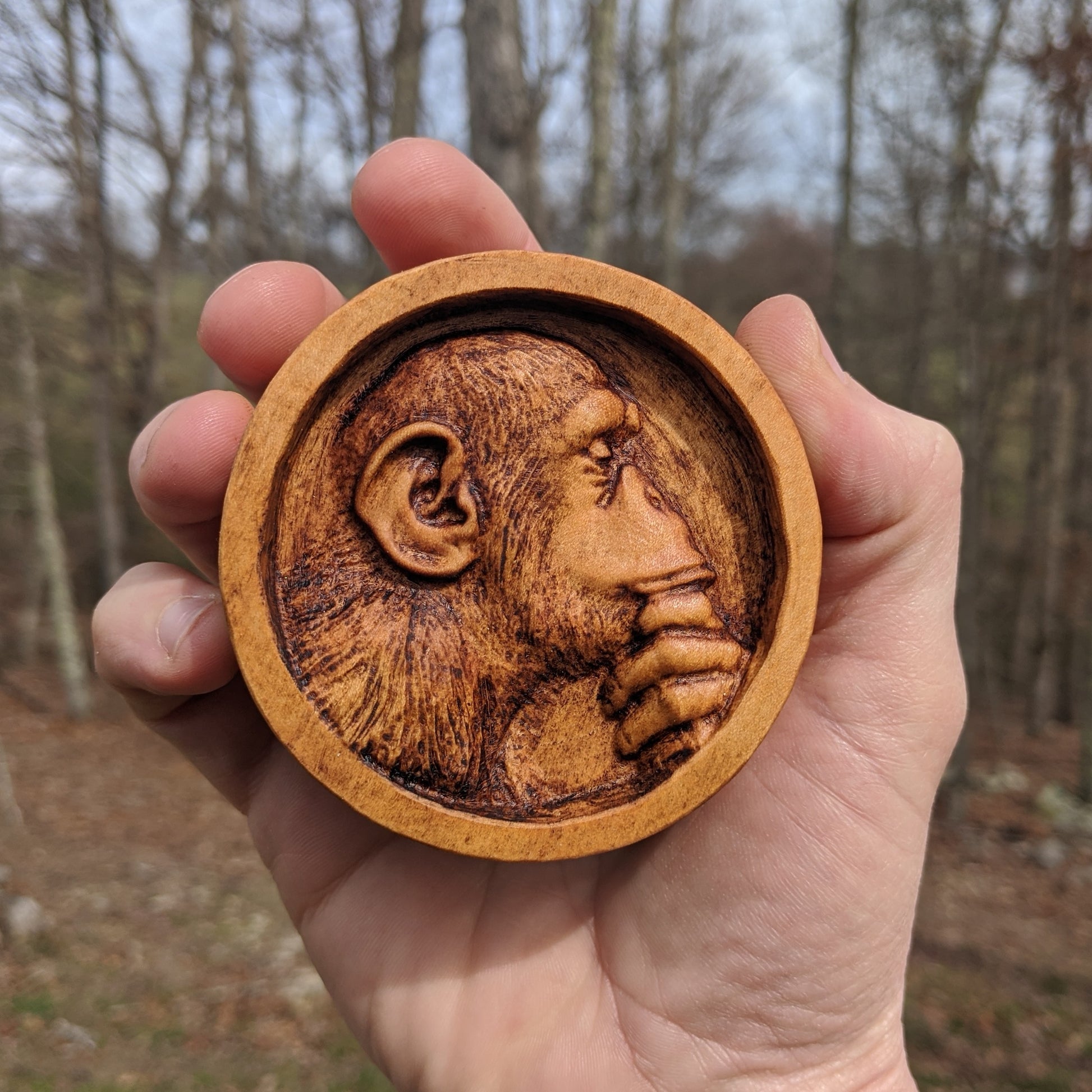 Round wooden box 3D carved with a recessed carving of a side profile view of an hairy ape rubbing his chin in his hand while in mid thought. Made from hard maple wood with a brown stain, being held in a hand against a forest background.