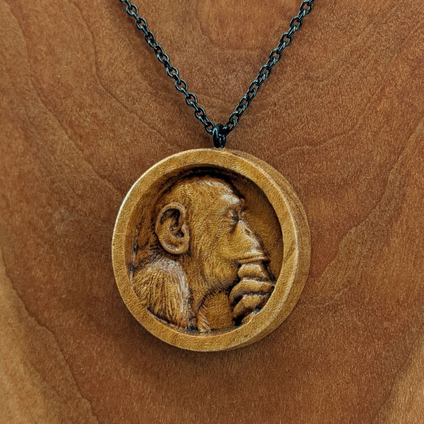 Wooden necklace pendant carved in the shape of a monkey with his hand on his chin. Made from hard maple wood and hanging from a black stainless steel chain against a cherry wood background.