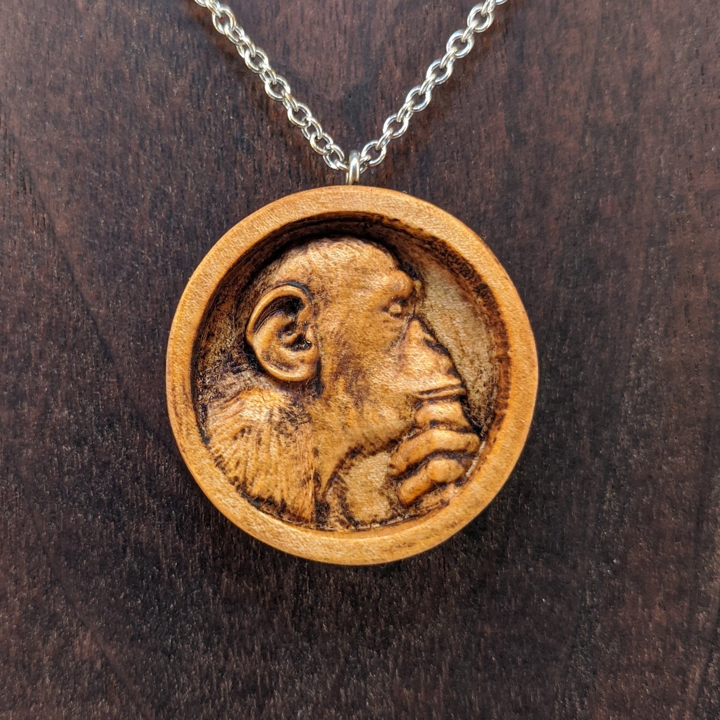 Wooden necklace pendant carved in the shape of a monkey with his hand on his chin. Made from hard maple wood and hanging from a silver stainless steel chain against a dark walnut background.