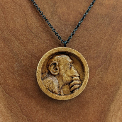 Wooden necklace pendant carved in the shape of a monkey with his hand on his chin. Made from hard maple wood and hanging from a black stainless steel chain against a cherry wood background.