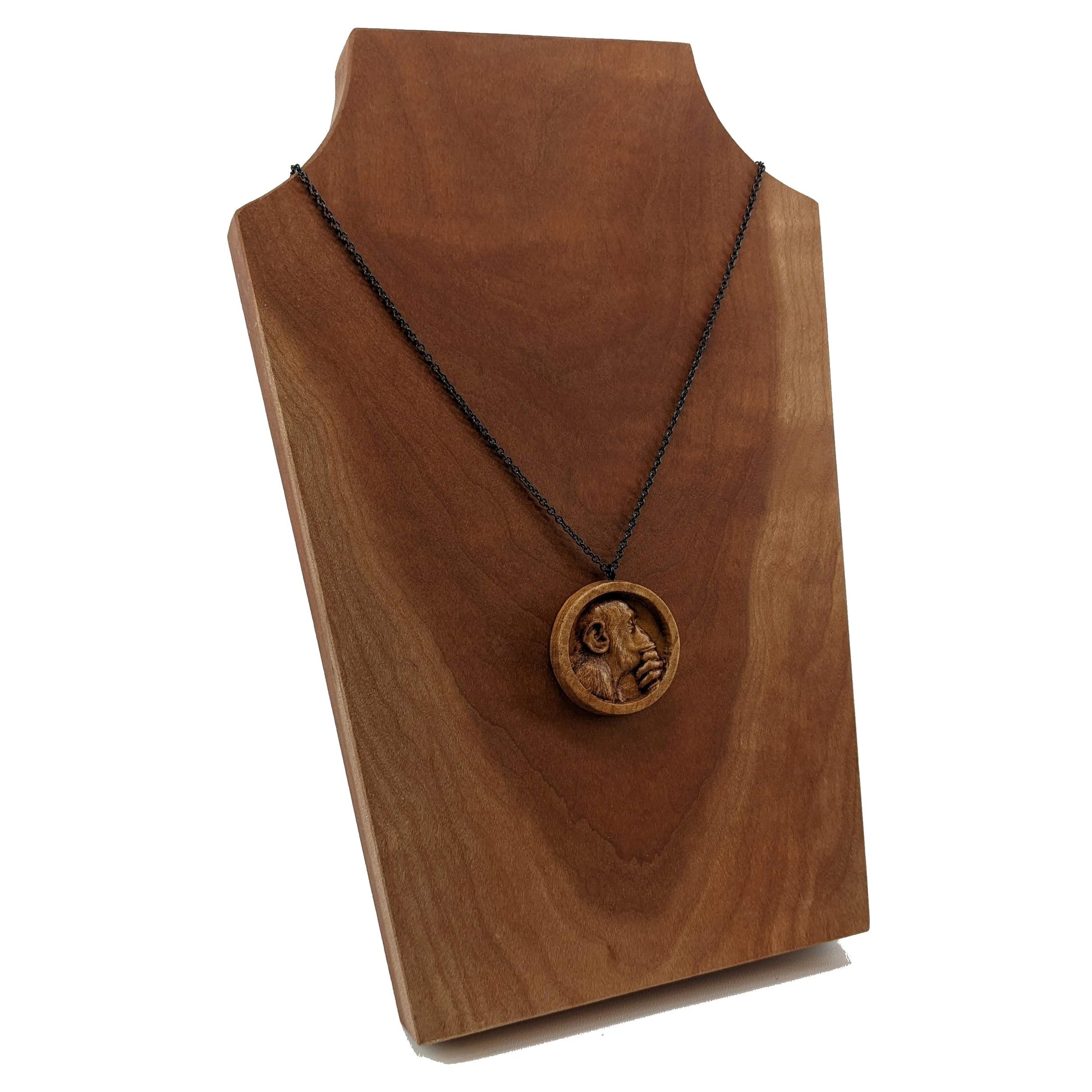 Wooden necklace pendant carved in the shape of a monkey with his hand on his chin. Made from hard maple wood and hanging from a black stainless steel chain against a cherry wood background.