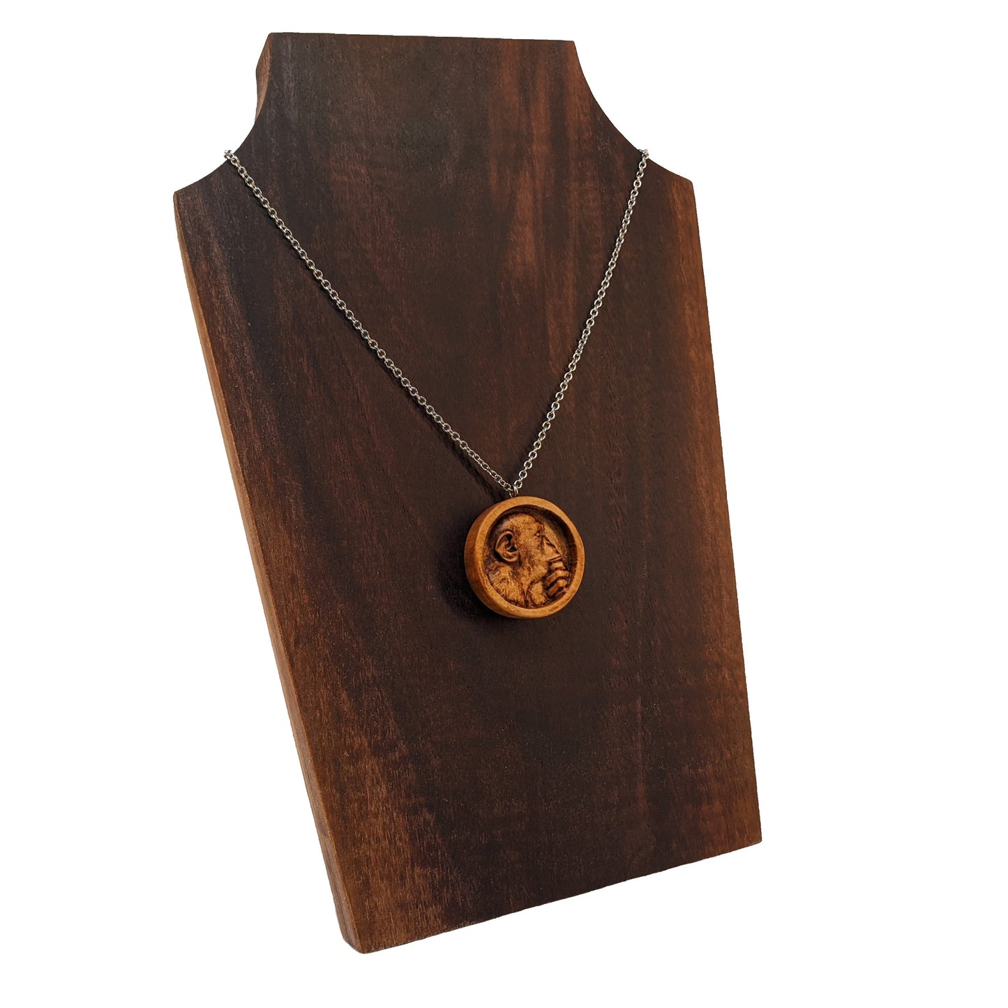 Wooden necklace pendant carved in the shape of a monkey with his hand on his chin. Made from hard maple wood and hanging from a silver stainless steel chain against a dark walnut display.