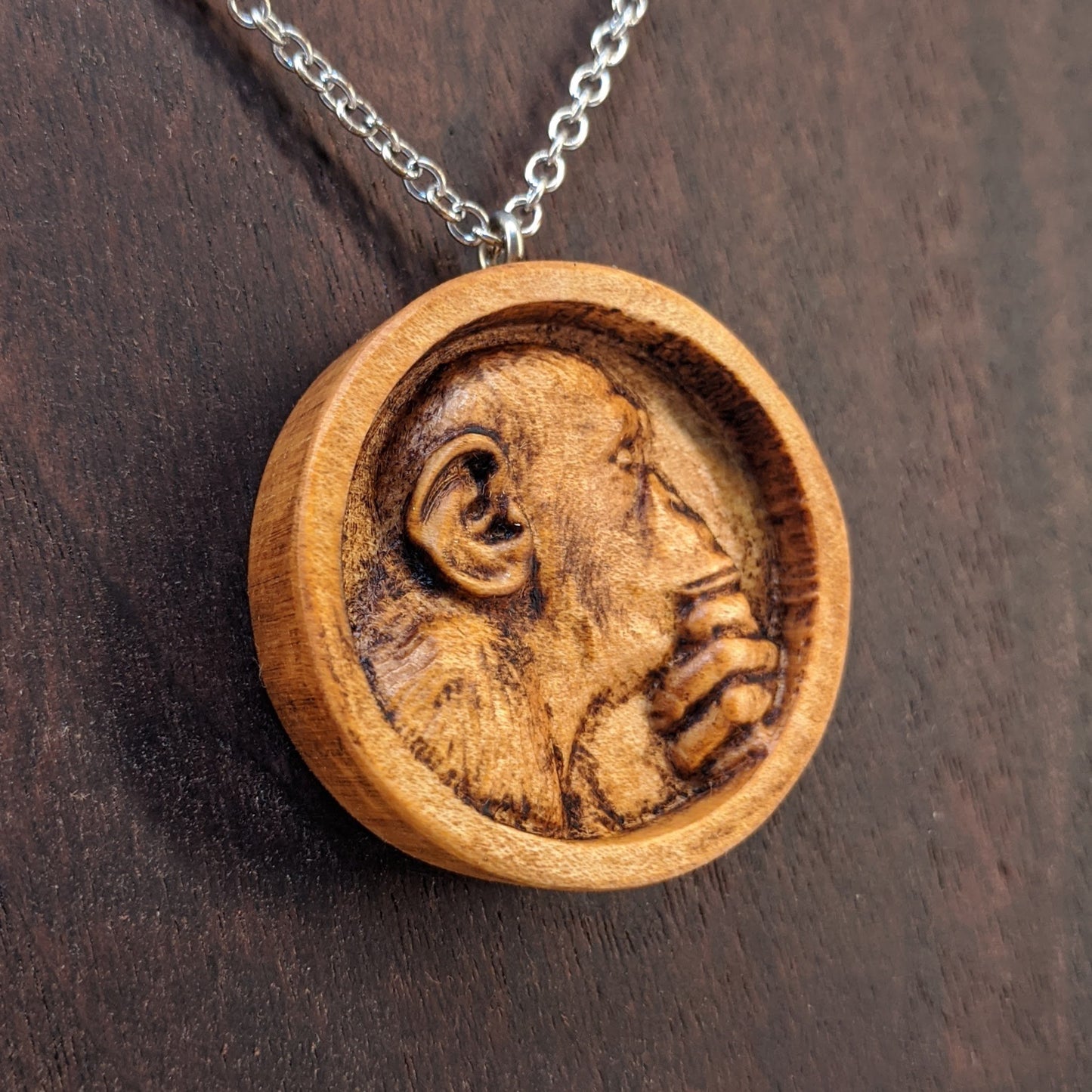 Wooden necklace pendant carved in the shape of a monkey with his hand on his chin. Made from hard maple wood and hanging from a silver stainless steel chain against a dark walnut background.