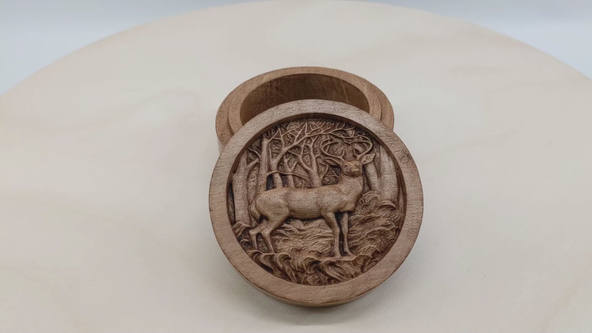 Round wooden box 3D carved with a buck deer standing in a forest. It stands in small grassy knolls in front of trees that have thin branches sticking out in every direction. Made from hard maple wood with a brown stain against a white background rotating on a table. 