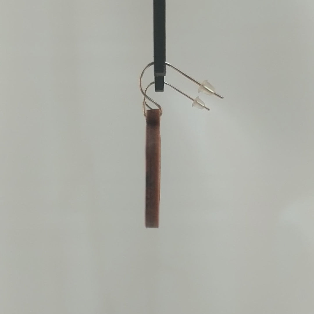 Pair of wooden earrings with silver stainless steel hooks. They are two toned light and dark brown miniature snare drums with drumsticks on top. Made from birch wood hanging and rotating against a white background.