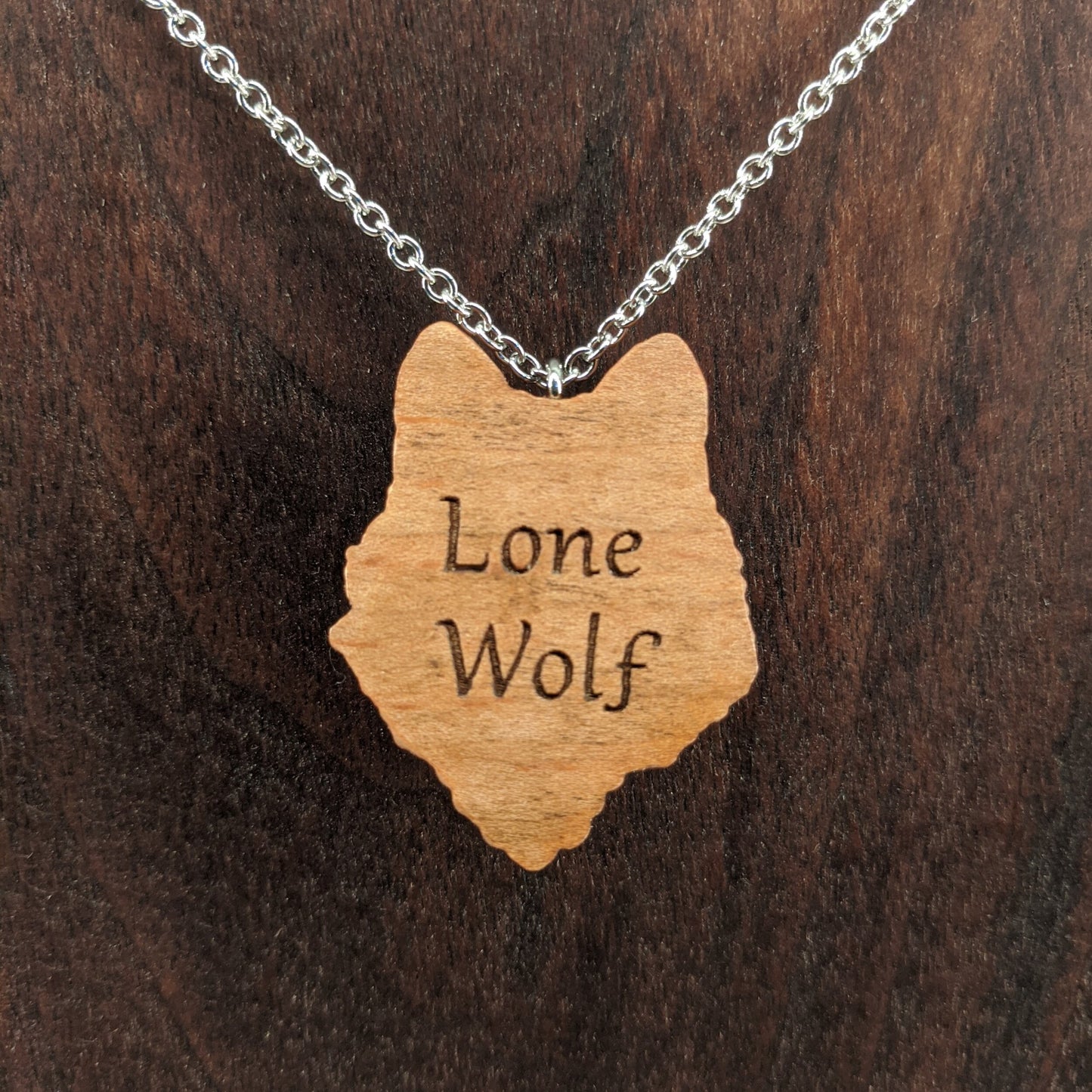 Wooden necklace pendant carved in the shaped of an wolf head. Made from hard maple wood. The backside is engraved with personalized wording. Hanging from a silver stainless steel chain against a dark walnut background.