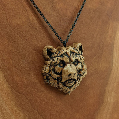 Wooden necklace pendant carved in the shape of a grizzly bears head. Made from hard maple wood. Hanging from a black stainless steel chain against a cherry wood background.