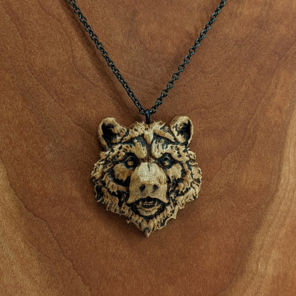 Wooden necklace pendant carved in the shape of a grizzly bears head. Made from hard maple wood. Hanging from a black stainless steel chain against a cherry wood background.