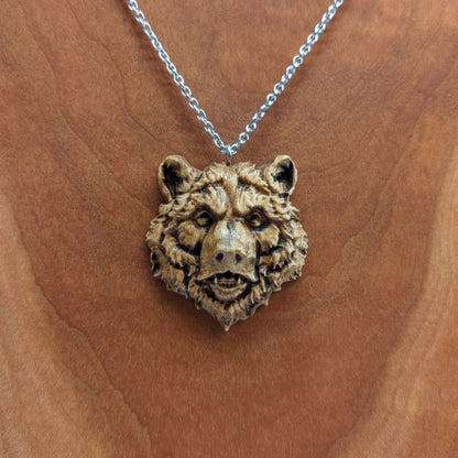 Wooden necklace pendant carved in the shape of a grizzly bears head. Made from hard maple wood. Hanging from a silver stainless steel chain against a cherry wood background.