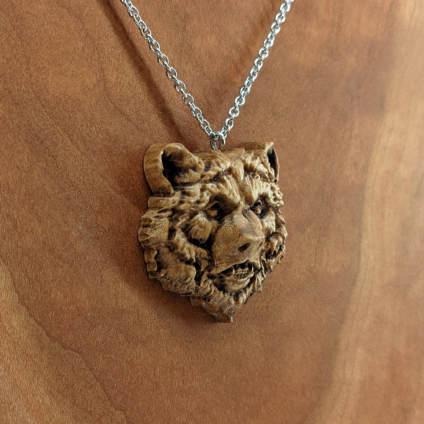 Wooden necklace pendant carved in the shape of a grizzly bears head. Made from hard maple wood. Hanging from a silver stainless steel chain against a cherry wood background.