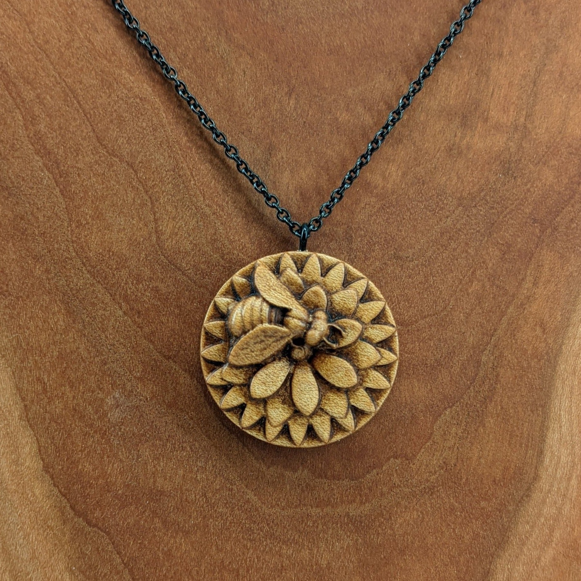 Wooden necklace pendant carved in the shape of a honey bee resting on a flower. Made from hard maple wood and hanging from a black stainless steel chain against a cherry wood background.