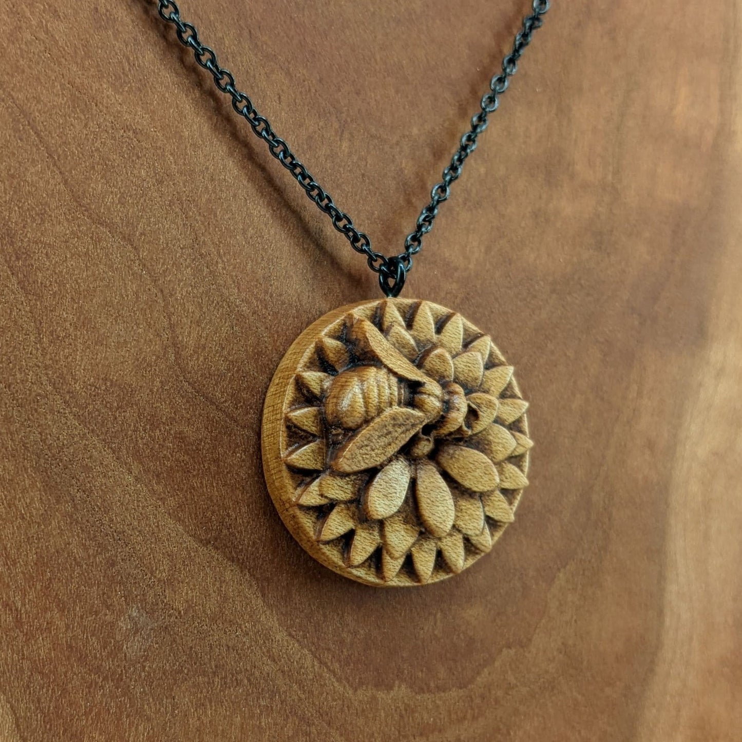 Wooden necklace pendant carved in the shape of a honey bee resting on a flower. Made from hard maple wood and hanging from a black stainless steel chain against a cherry wood background.