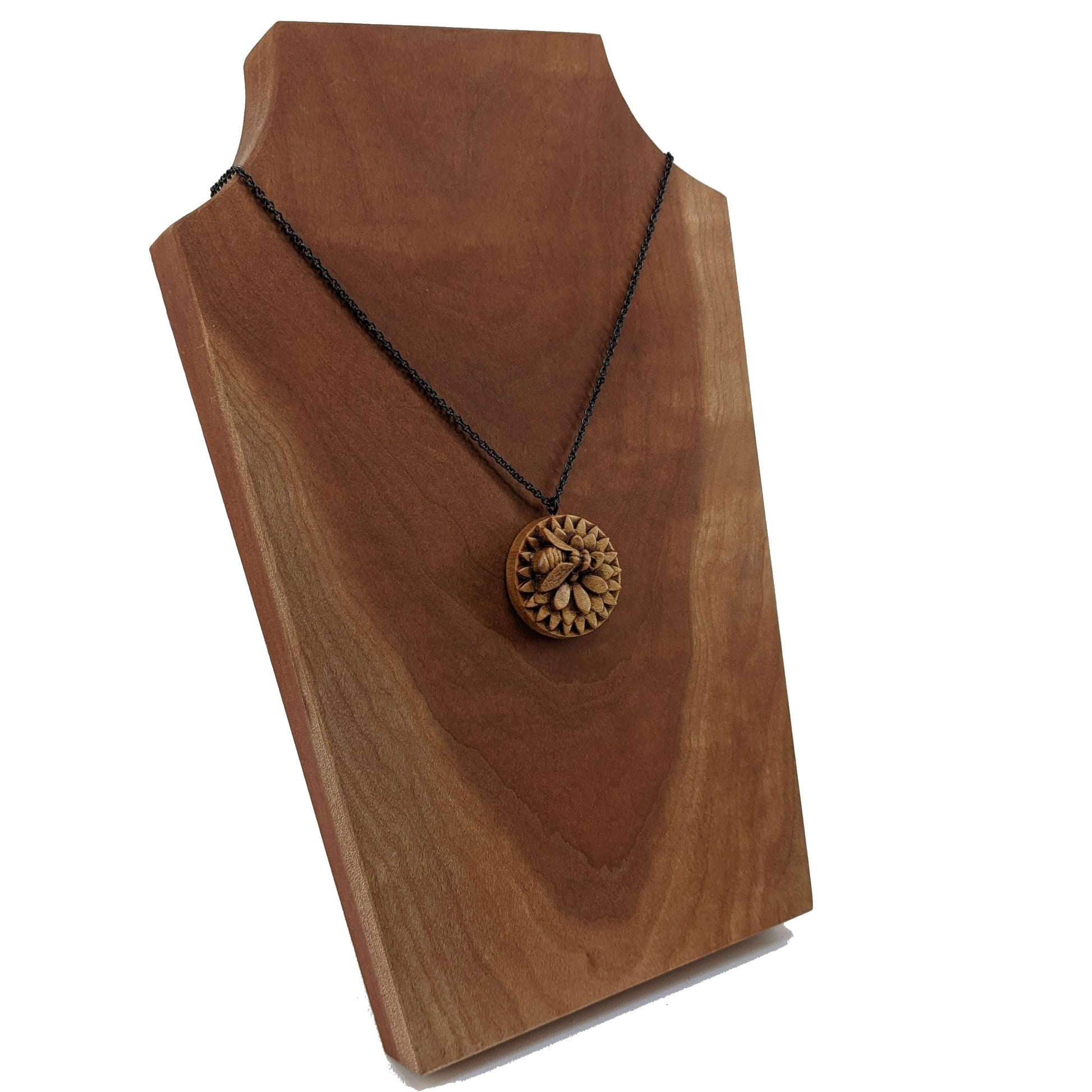 Wooden necklace pendant carved in the shape of a honey bee resting on a flower. Made from hard maple wood and hanging from a black stainless steel chain against a cherry wood display..