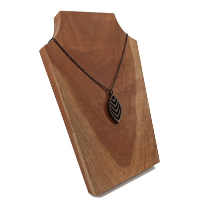 Wooden necklace pendant cut into a teardrop shape with a chevron pattern cut throughout it. Made from walnut and hanging from a black stainless steel chain against a cherry wood background.