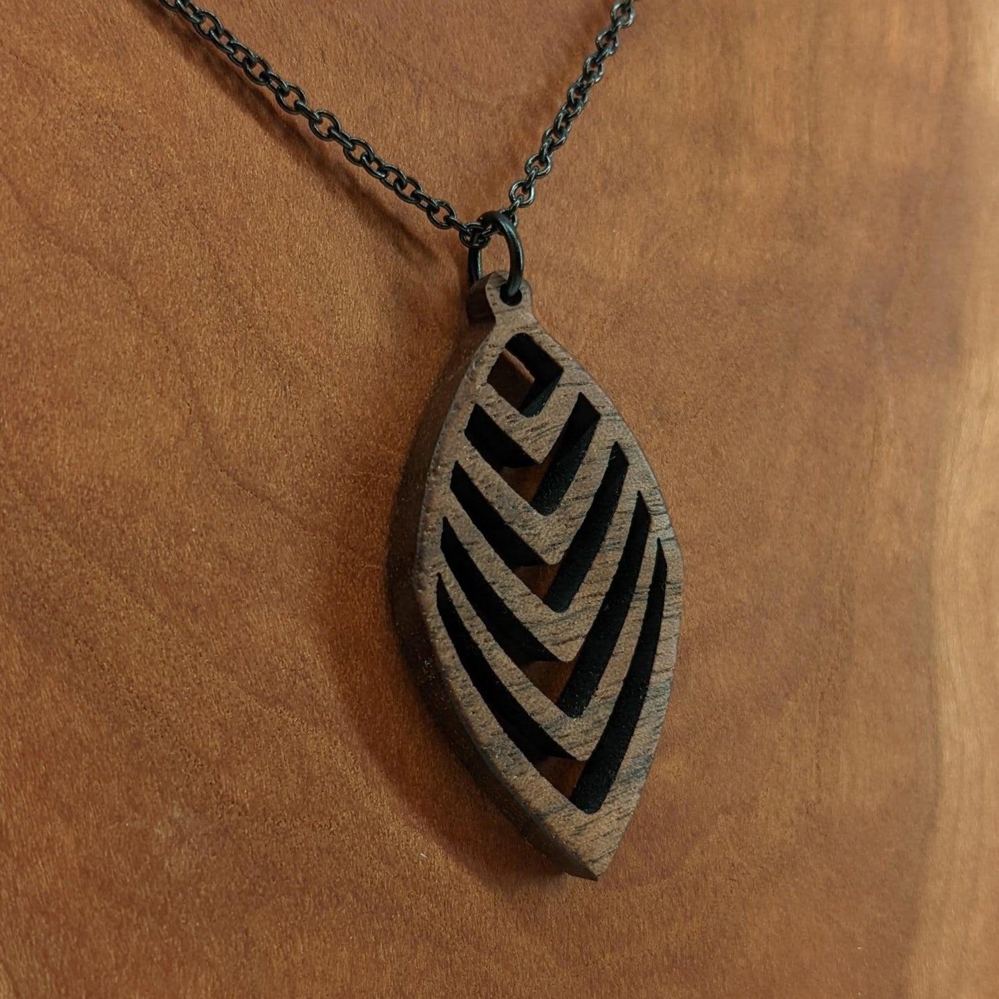 Wooden necklace pendant cut into a teardrop shape with a chevron pattern cut throughout it. Made from walnut and hanging from a black stainless steel chain against a cherry wood background.