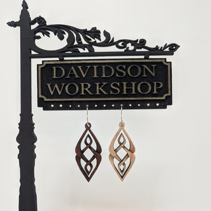 Pair of wooden earrings with silver stainless steel hooks. They are diamond shaped woven patterned carvings with inner diamonds woven within. Made from hard maple and walnut wood hanging from a model Davidson Workshop sign against a white background.