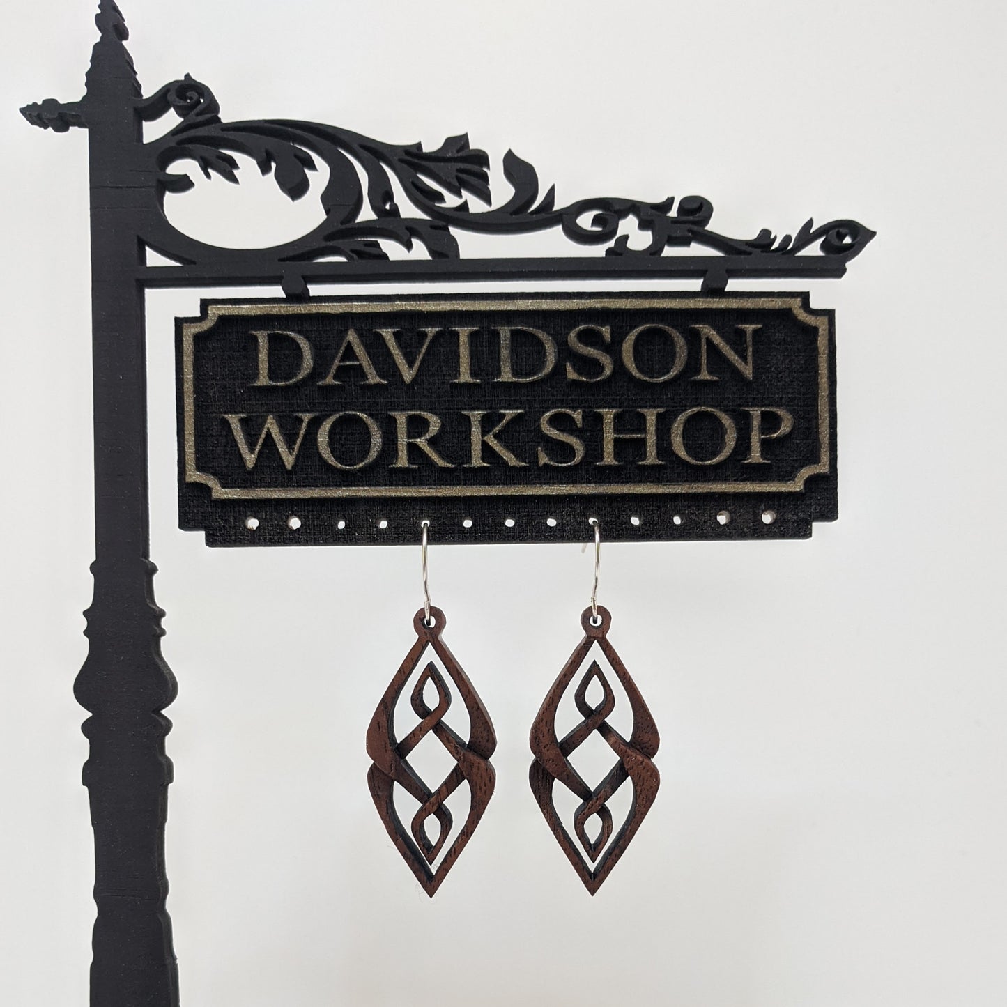 Pair of wooden earrings with silver stainless steel hooks. They are diamond shaped woven patterned carvings with inner diamonds woven within. Made from dark walnut hanging from a model Davidson Workshop sign against a white background.