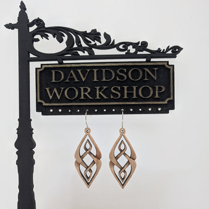 Pair of wooden earrings with silver stainless steel hooks. They are diamond shaped woven patterned carvings with inner diamonds woven within. Made from hard maple hanging from a model Davidson Workshop sign against a white background. (Close up View)