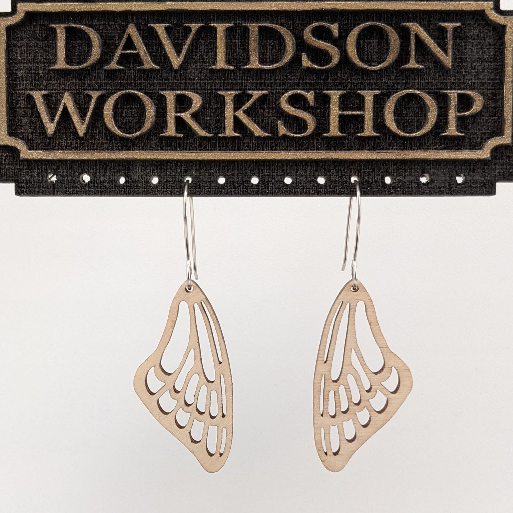 Pair of wooden earrings with silver stainless steel hooks. They are a pale natural finish cutout of a butterflies wing hanging downward. Made from birch wood hanging from a model Davidson Workshop sign against a white background. (Close up View)