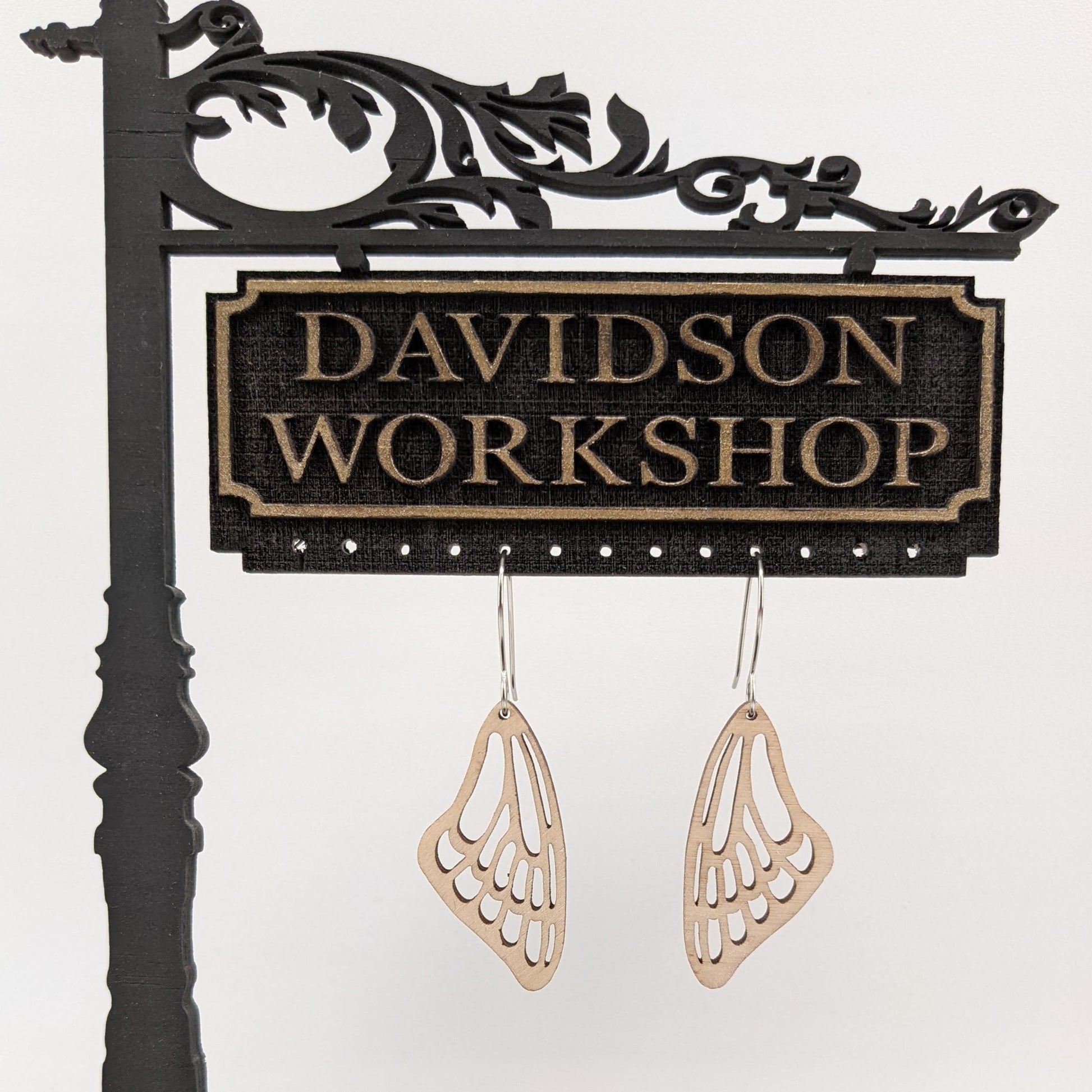 Pair of wooden earrings with silver stainless steel hooks. They are a pale natural finish cutout of a butterflies wing hanging downward. Made from birch wood hanging from a model Davidson Workshop sign against a white background.