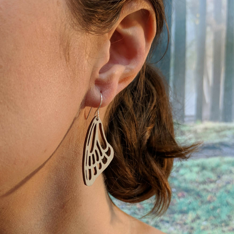 Pair of wooden earrings with silver stainless steel hooks. They are a pale natural finish cutout of a butterflies wing hanging downward. Made from birch wood, being modeled by a women in front of a nature scene.