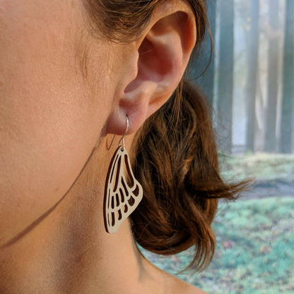 Pair of wooden earrings with silver stainless steel hooks. They are a pale natural finish cutout of a butterflies wing hanging downward. Made from birch wood, being modeled by a women in front of a nature scene.