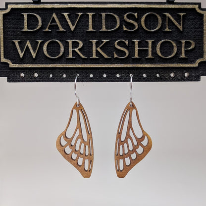 Pair of wooden earrings with silver stainless steel hooks. They are a pale natural finish cutout of a butterflies wing hanging downward. Made from birch wood hanging from a model Davidson Workshop sign against a white background. (Close up View)