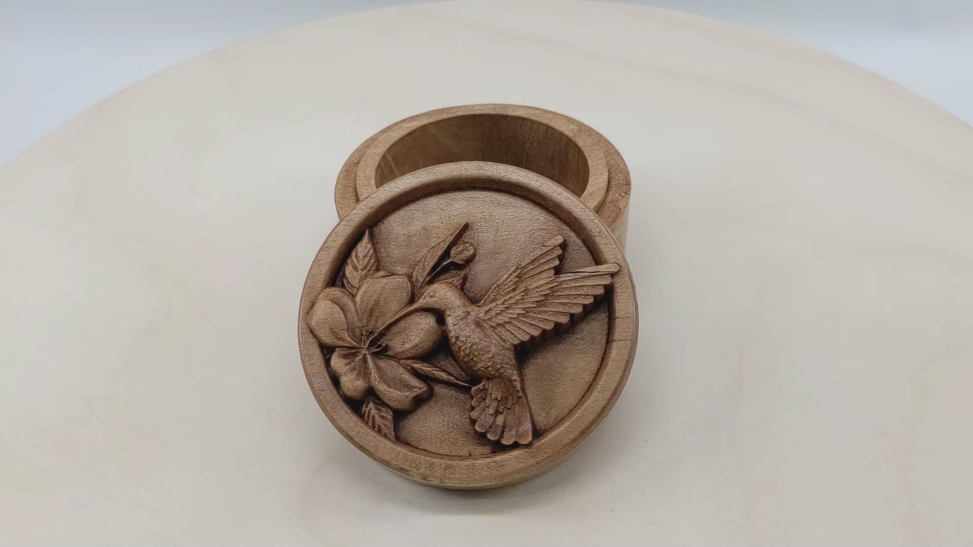 Round wooden box 3D carved with a recessed hummingbird hovering and drinking nectar from a flower. It is facing to the left with its delicate wings aflutter as it leans in to drink. Made from hard maple wood stained brown against a white background rotating on a table.