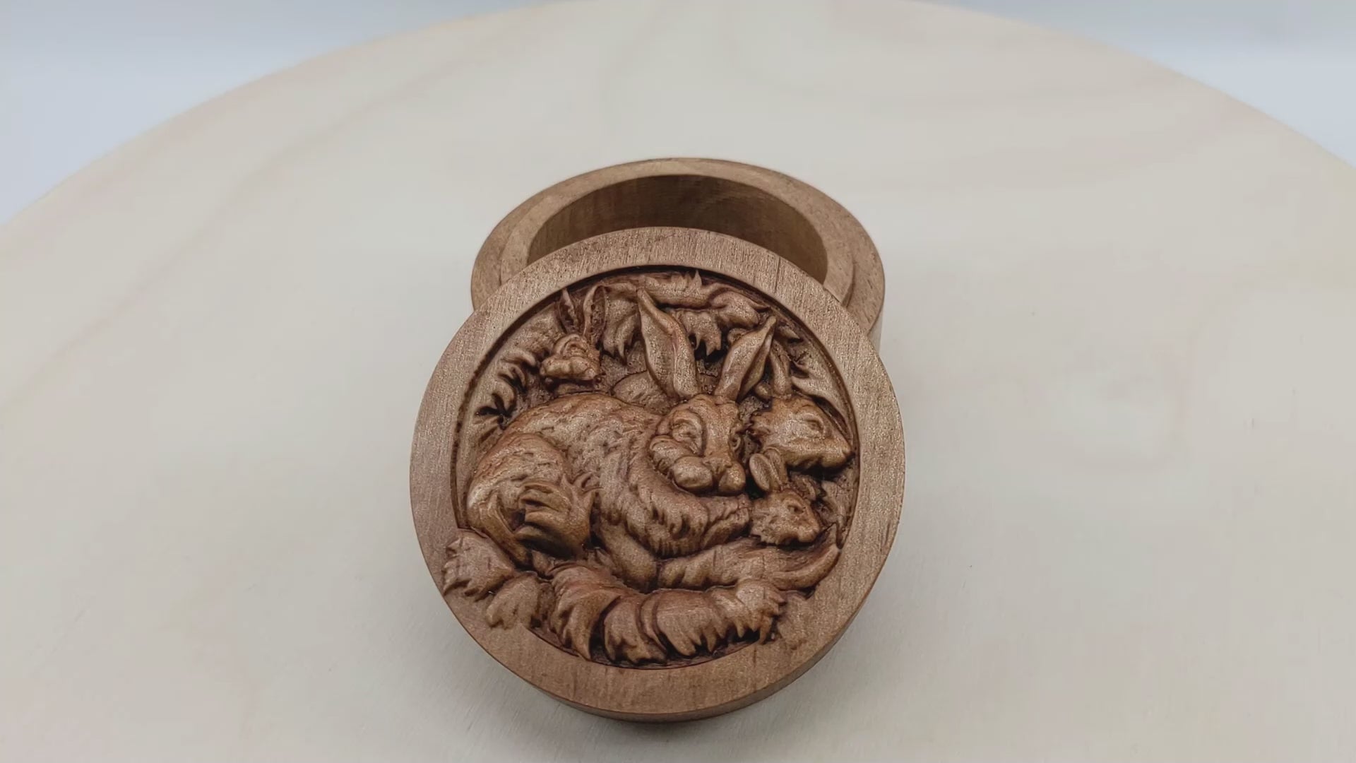 Round wooden box 3D carved with a family of rabbits together in their nest. Mother rabbit has bushy fur, tall ears and heads of three baby rabbits peaking out from behind her. Made from hard maple wood stained brown against a white background rotating on a table.