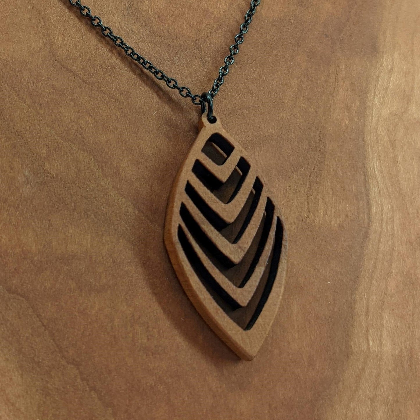 Wooden necklace pendant cut into a teardrop shape with a chevron pattern cut throughout it. Made from cherry and hanging from a black stainless steel chain against a cherry background.