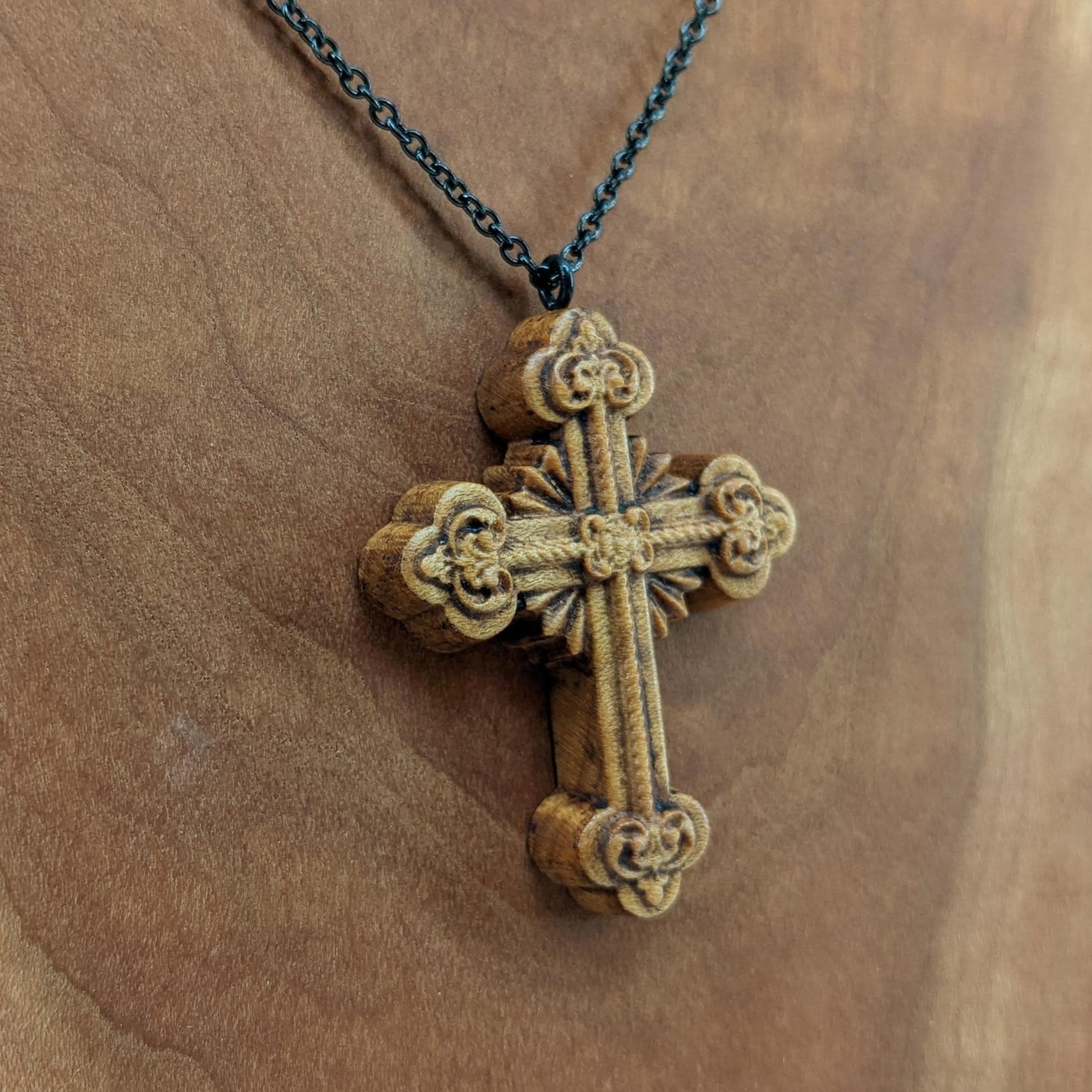 Wooden necklace pendant carved in the shape of an ornate cross. Made from hard maple wood and hanging from a black stainless steel chain against a cherry wood background.