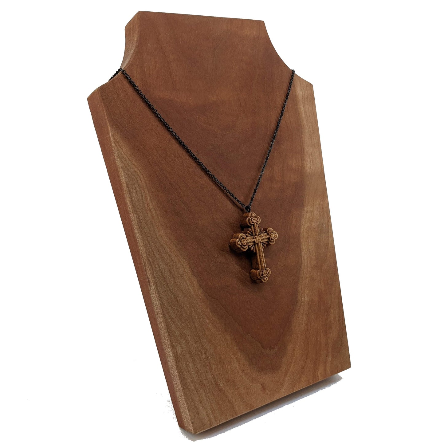 Wooden necklace pendant carved in the shape of an ornate cross. Made from hard maple wood and hanging from a black stainless steel chain against a cherry wood background.