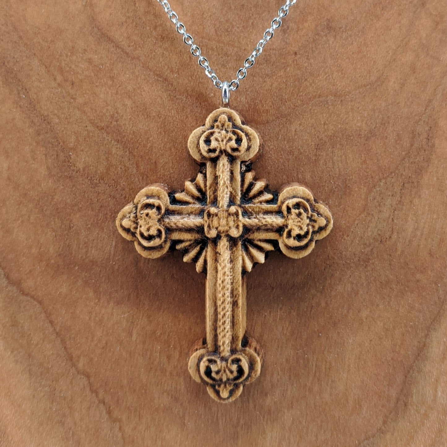 Wooden necklace pendant carved in the shape of an ornate cross. Made from hard maple wood and hanging from a silver stainless steel chain against a cherry wood background.