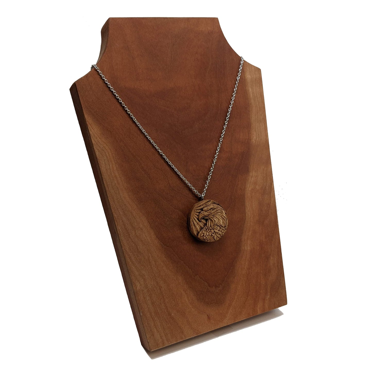 Round wooden necklace pendant carved in the shape of an eagles head. Made from hard maple wood. Hanging from a silver stainless steel chain against a cherry wood background.