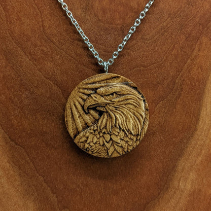 Round wooden necklace pendant carved in the shape of an eagles head. Made from hard maple wood. Hanging from a silver stainless steel chain against a cherry wood background.