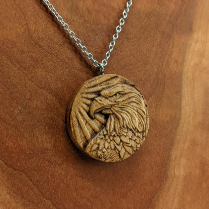 Round wooden necklace pendant carved in the shape of an eagles head. Made from hard maple wood. Hanging from a silver stainless steel chain against a cherry wood background.