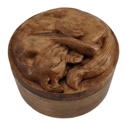 Round wooden box 3D carved with a short haired cat with textured fur curled up into a ball sleeping. It has a peaceful rested expression on its face and legs all crossed together. Made from hard maple wood stained brown against a white background.