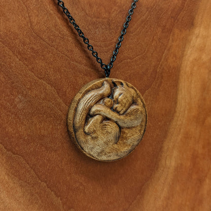 Wooden necklace pendant carved into the shape of a cat curled into a round ball. Hanging from a black stainless steel chain against a cherry wood background.