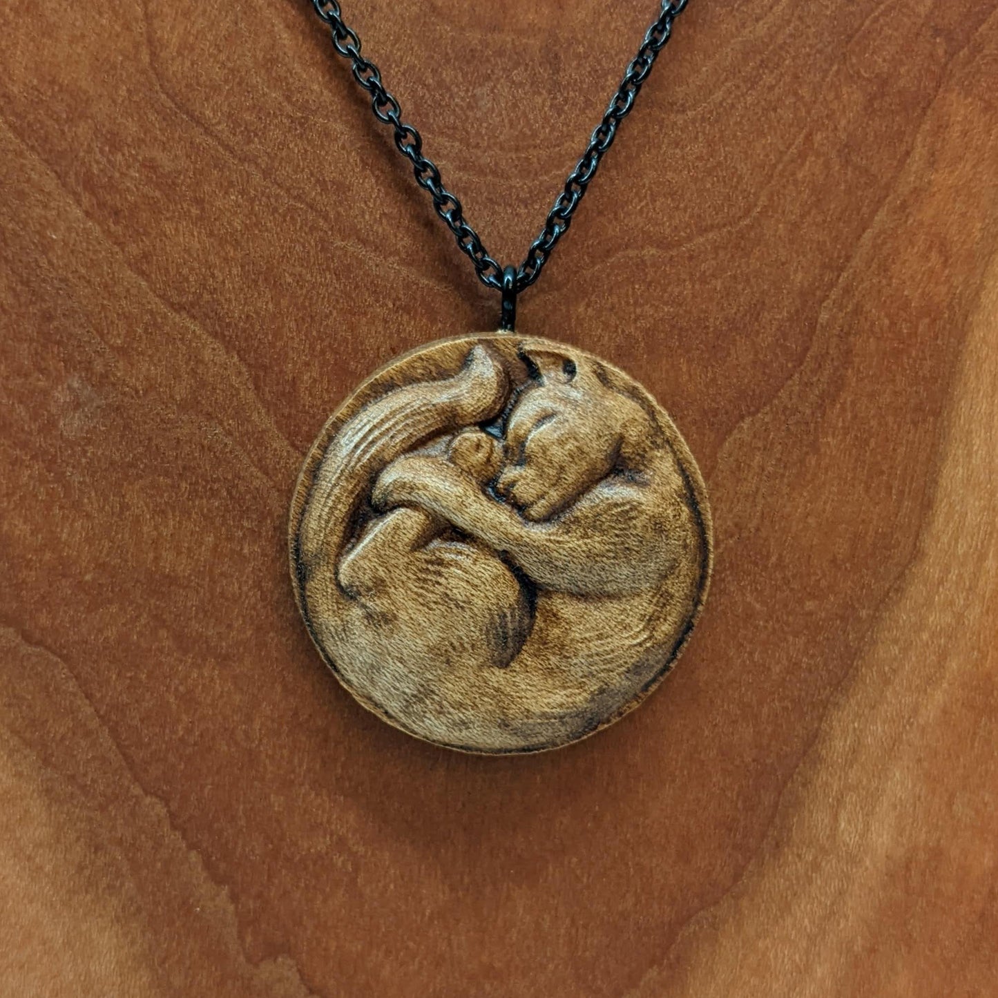 Wooden necklace pendant carved into the shape of a cat curled into a round ball. Hanging from a black stainless steel chain against a cherry wood background.